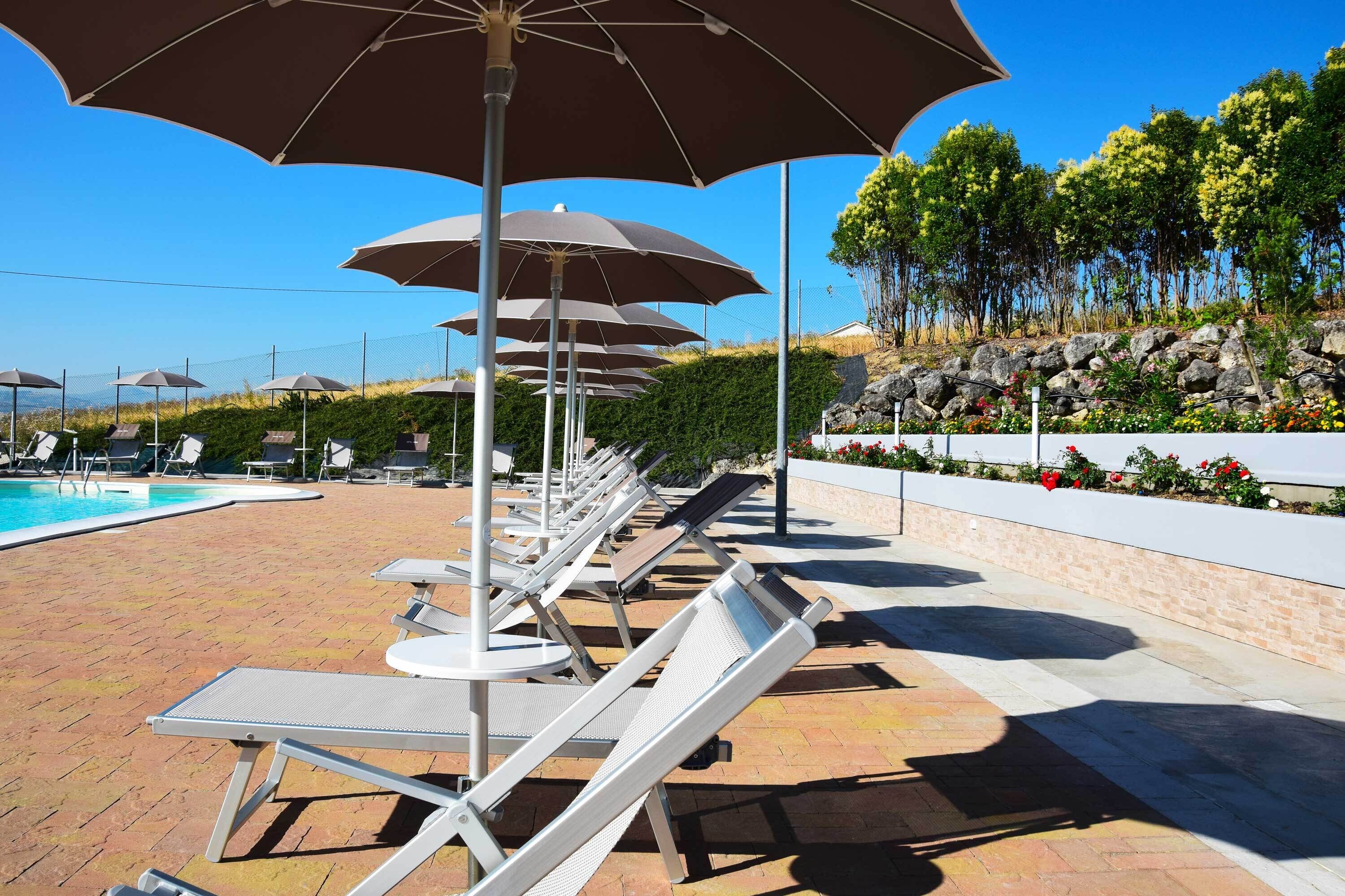 Uma área à beira da piscina com espreguiçadeiras e guarda-sóis, céu claro e um jardim com pedras e plantas.