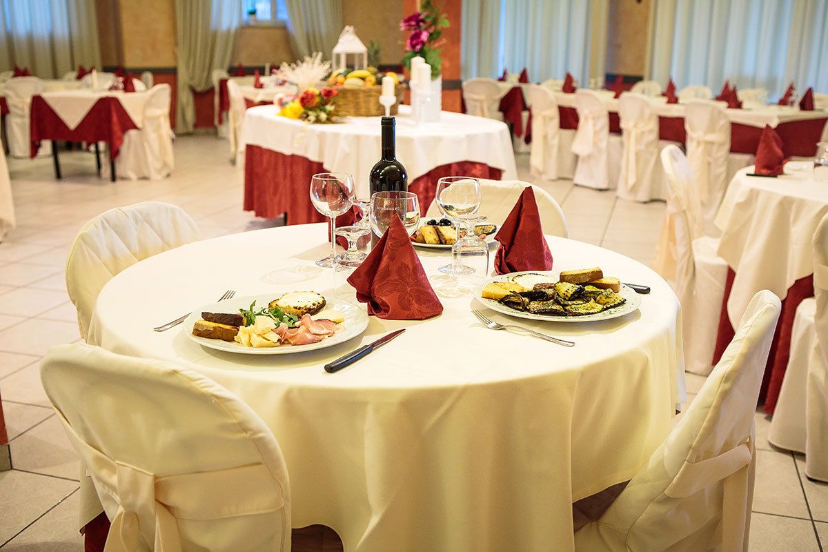A banquet hall with round tables set for a formal event, each with white tablecloths, red napkins, and a bottle of wine.