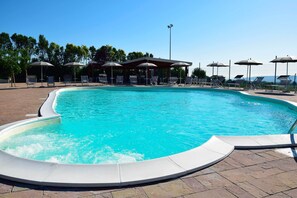 Una piscina con sillas de descanso y sombrillas, rodeada por un patio de ladrillo y árboles.