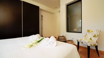 Una cama bien hecha con toallas enrolladas y un ramo de flores, una silla con estampado de hojas, y un ventanal grande en una habitación de hotel.