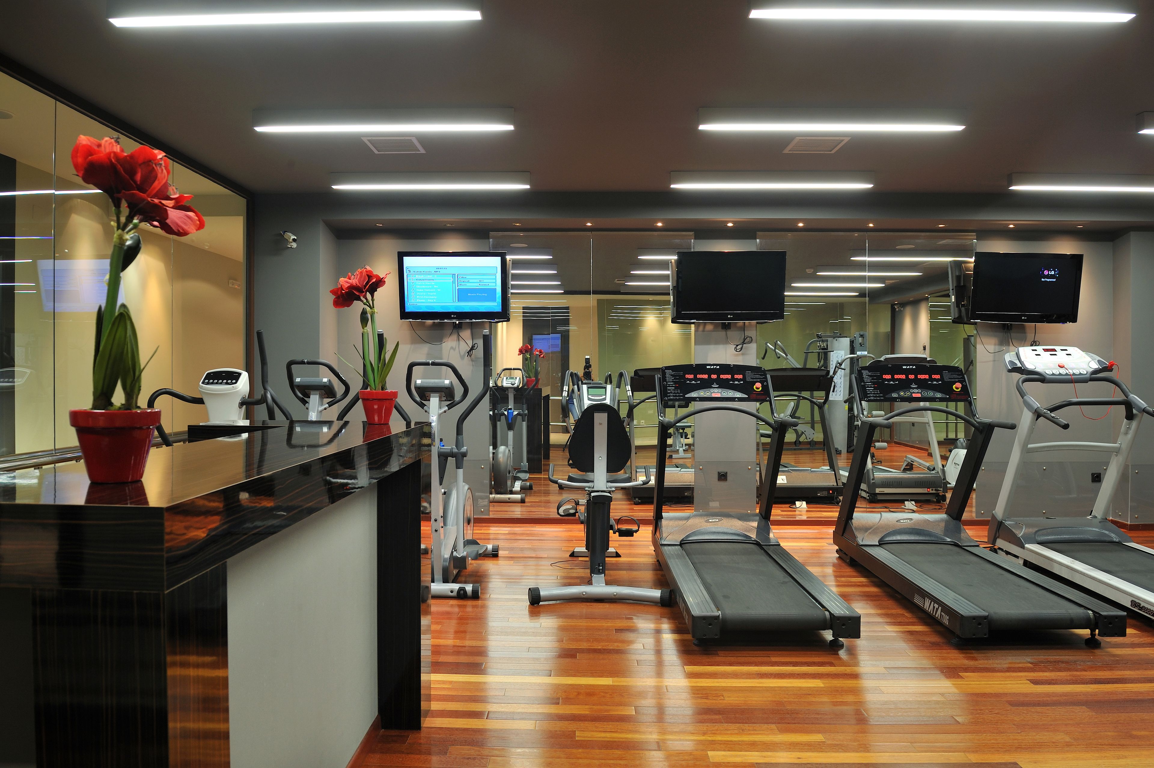 Une salle de sport moderne équipée de machines de cardio, de postes de musculation et d’un espace d’accueil avec un comptoir et des plantes en pot.