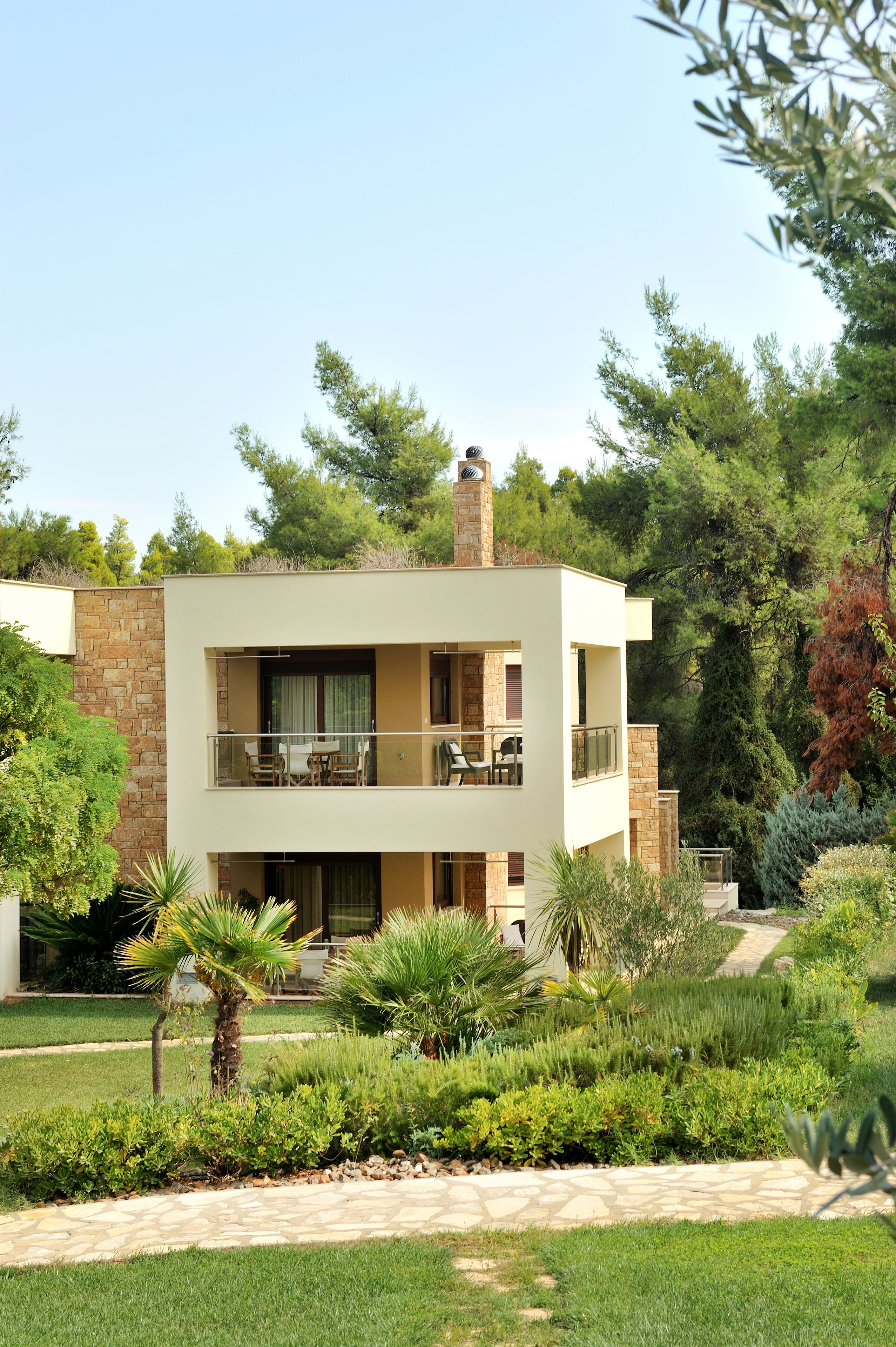 A modern two-story building with a balcony, surrounded by lush greenery and a stone pathway.