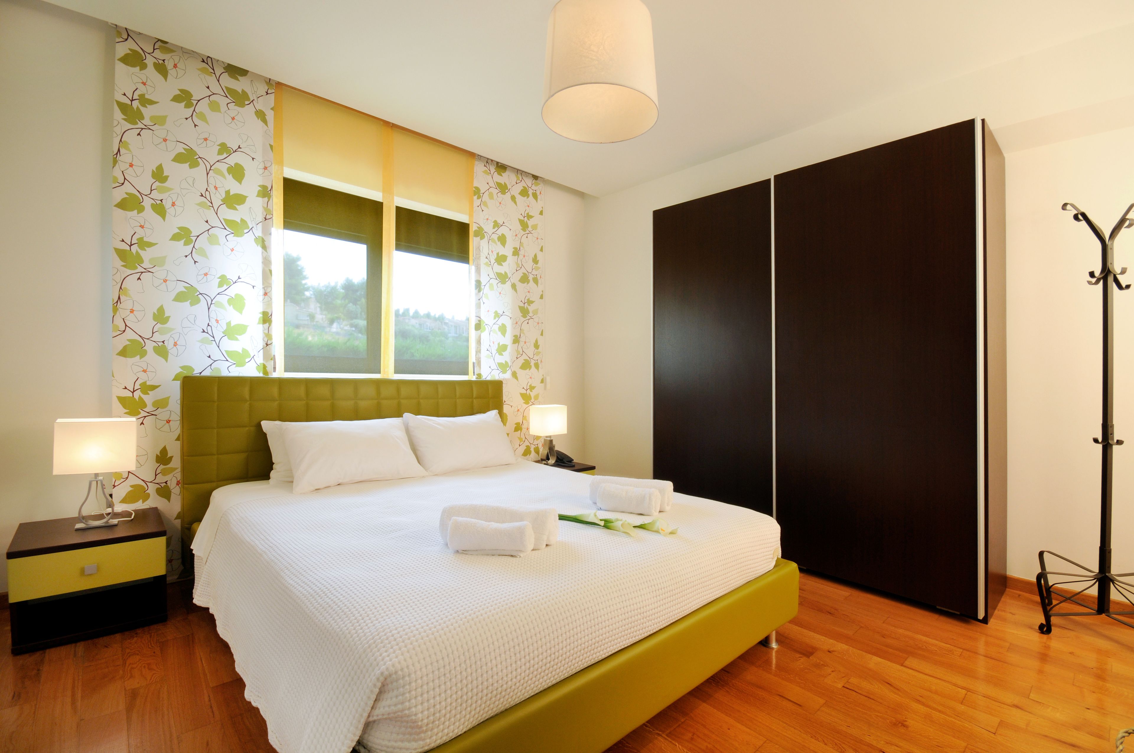 A neatly made bed with white linens, a green headboard, and two bedside lamps. There is a large wardrobe and a coat rack to the right.