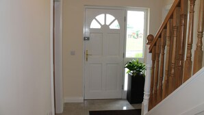 A hallway leading to a white door with a semi-circular window, a black potted plant, and a wooden staircase with a white handrail. - Quay Rest  (Rooskey)