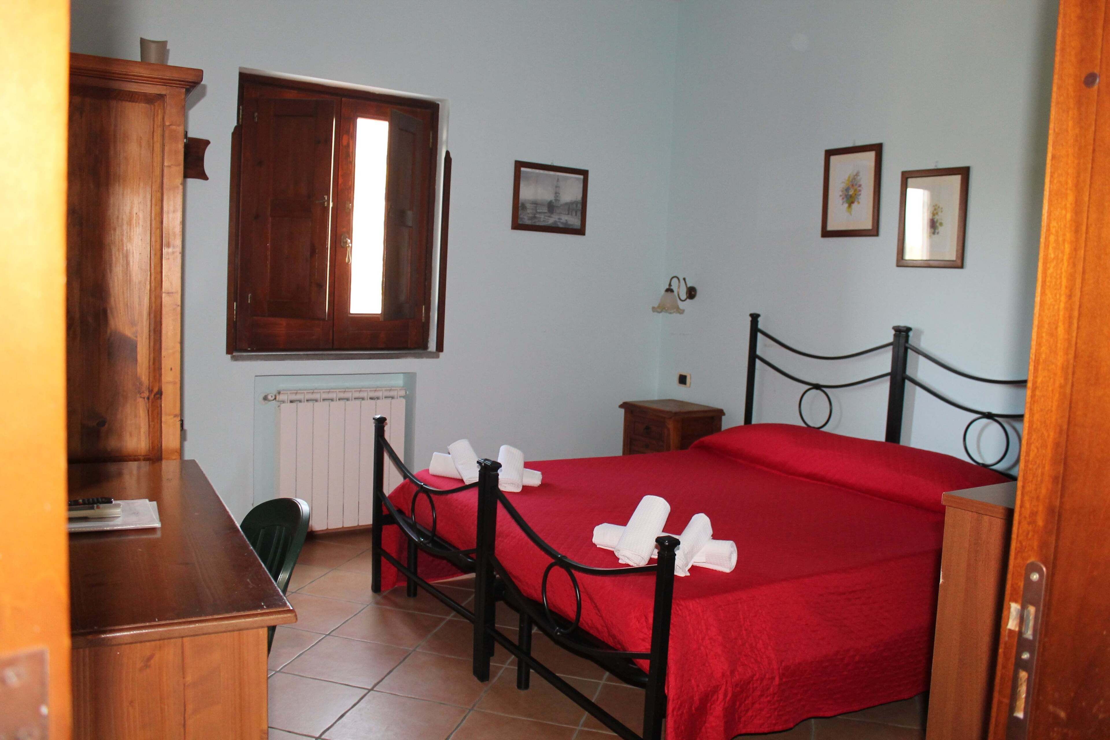 Un dormitorio con estructura de cama de madera, colcha roja, escritorio de madera y una silla.