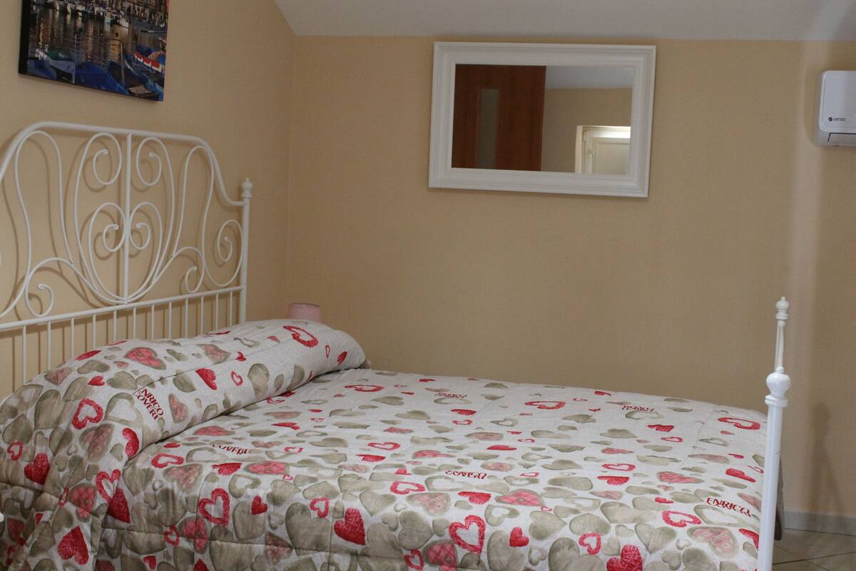 Une chambre à coucher avec un lit, une tête de lit en métal blanche, un mur de couleur beige, un miroir avec un cadre blanc et un petit appareil de climatisation.