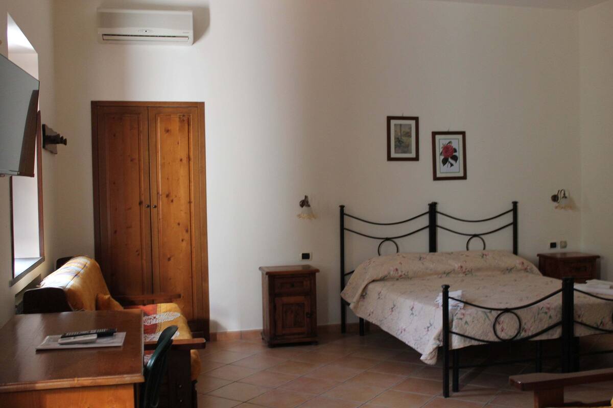 Une chambre à coucher avec une armoire en bois, un lit recouvert d’une couvre-lit à motifs floraux, une table de chevet et un petit meuble.