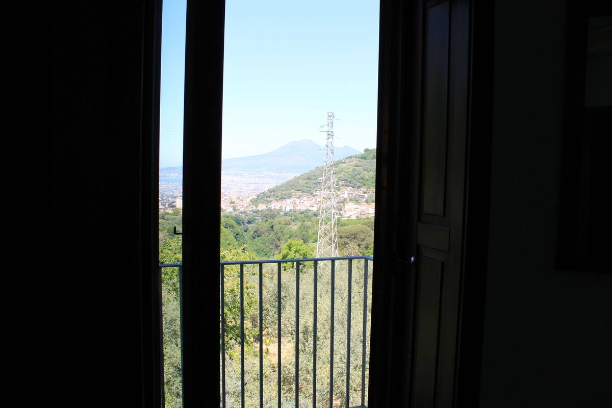 Une vue depuis un balcon doté d’une rambarde en métal, donnant sur un paysage urbain avec des bâtiments et une montagne à l’horizon.