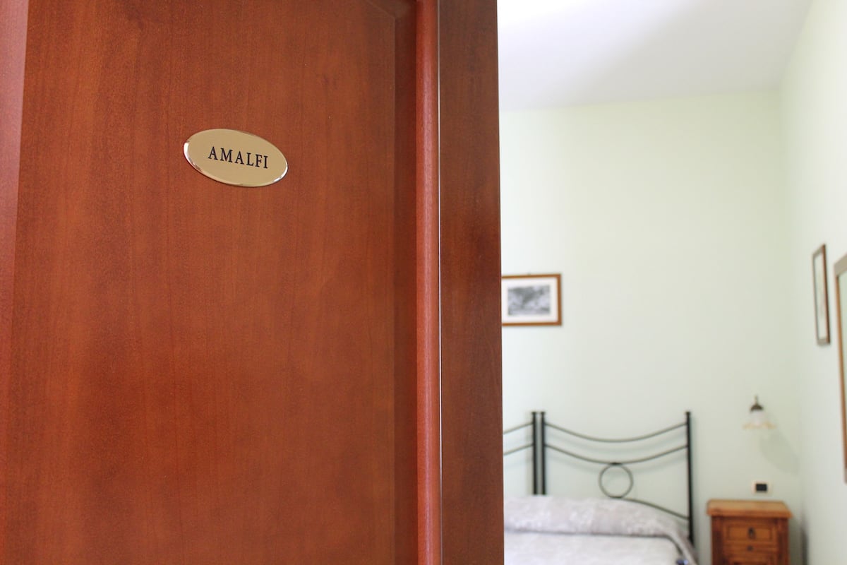 Une porte en bois avec une plaque portant l’inscription « AMALFI » et une chambre à coucher comprenant un lit, une table de chevet et une lampe fixée au mur.