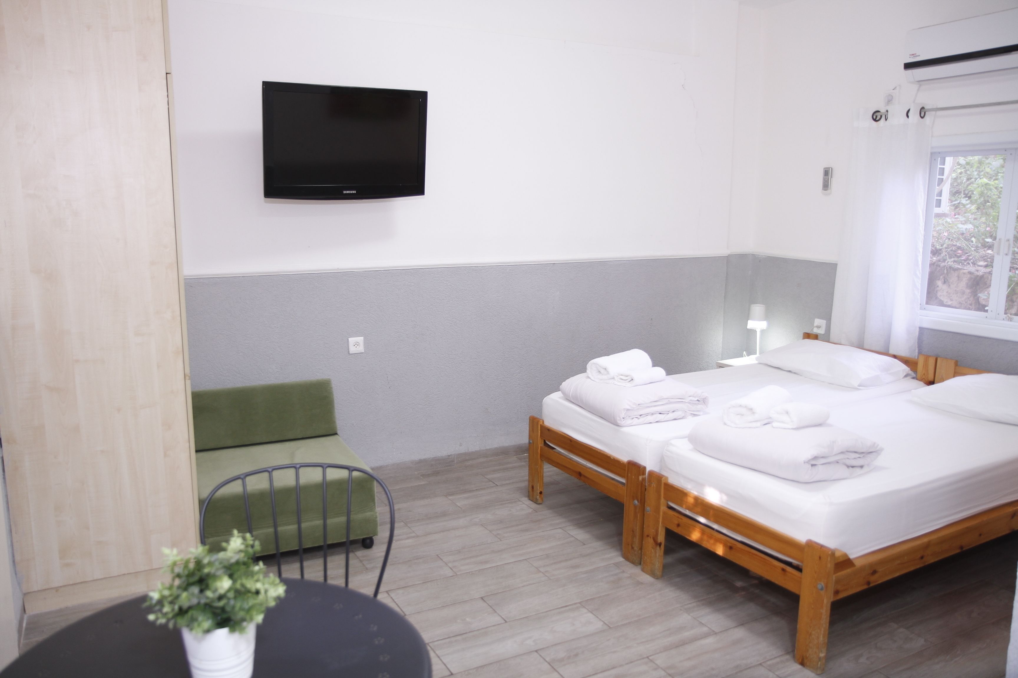 Ein Hotelzimmer mit Bett, einem an der Wand befestigten Fernseher, einem kleinen Tisch mit Pflanze und einem mit Vorhängen versehenen Fenster.