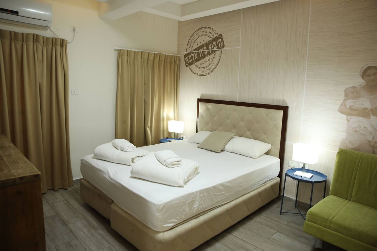A hotel room with a large bed, two bedside tables, a green armchair, and a wall-mounted air conditioning unit.