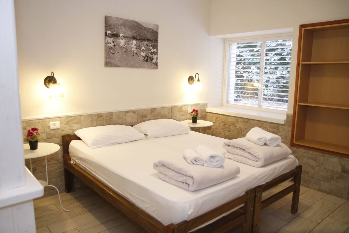 A neatly made bed with white linens and towels, a small round table with a potted plant, and a window with blinds.