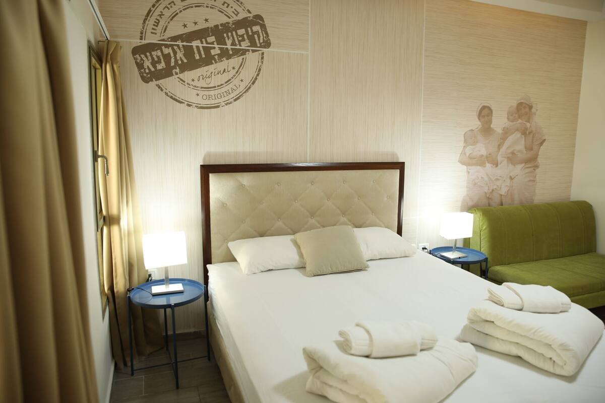 A hotel room with a large bed, two bedside tables, a green armchair, and a wall with a circular design.