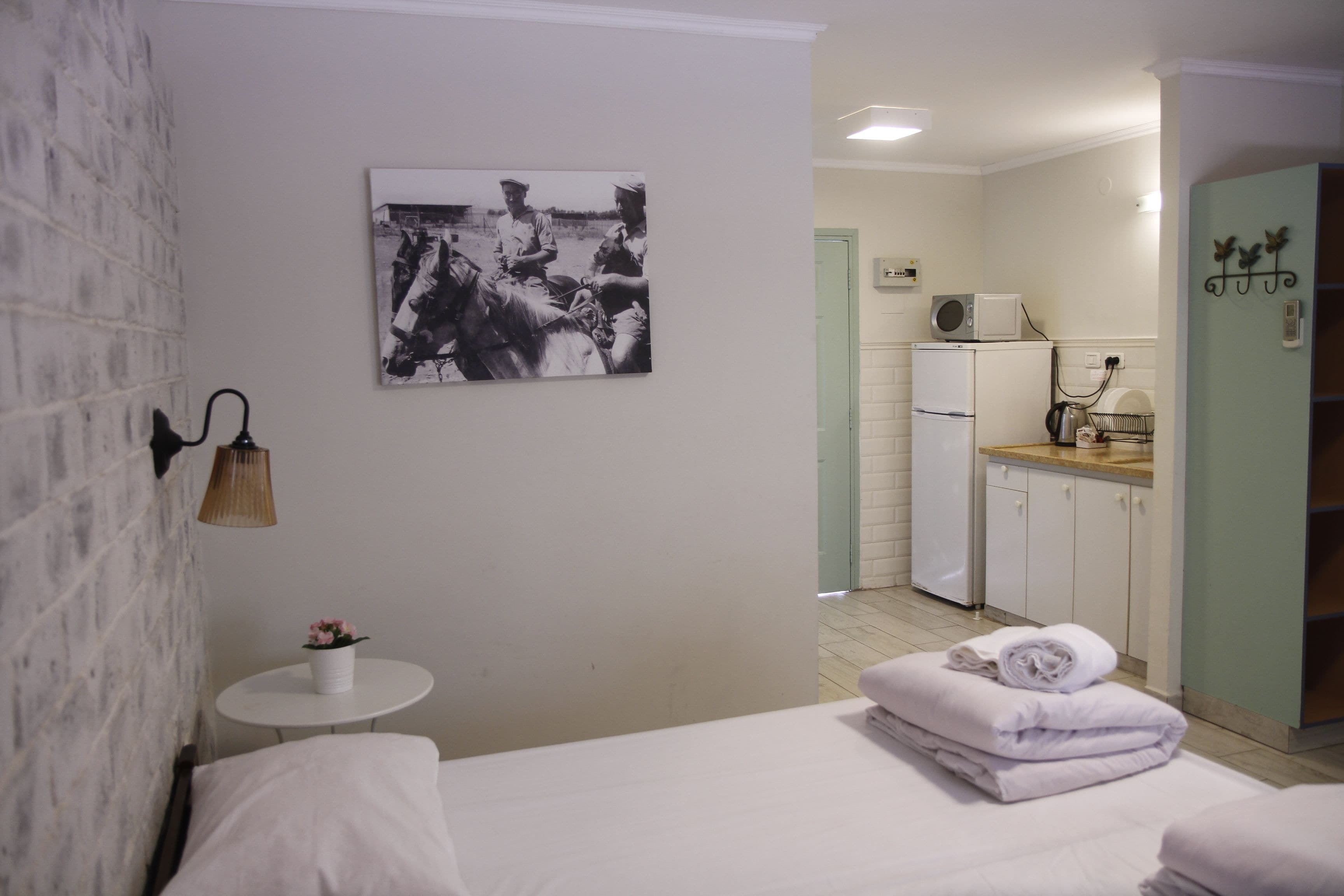 A hotel room with a bed, a small round table with a plant, a wall-mounted lamp, a picture of people on horseback, and a kitchenette in the background.