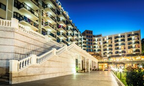 A multi-story hotel with a grand staircase leading to the entrance, multiple balconies, and a well-lit lobby area.