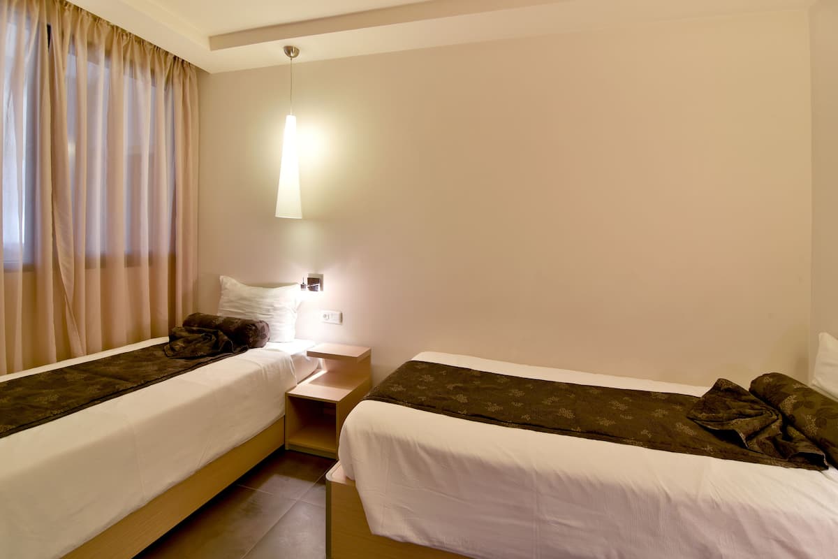 A hotel room with two single beds, a bedside table, a lamp, and a window with curtains.
