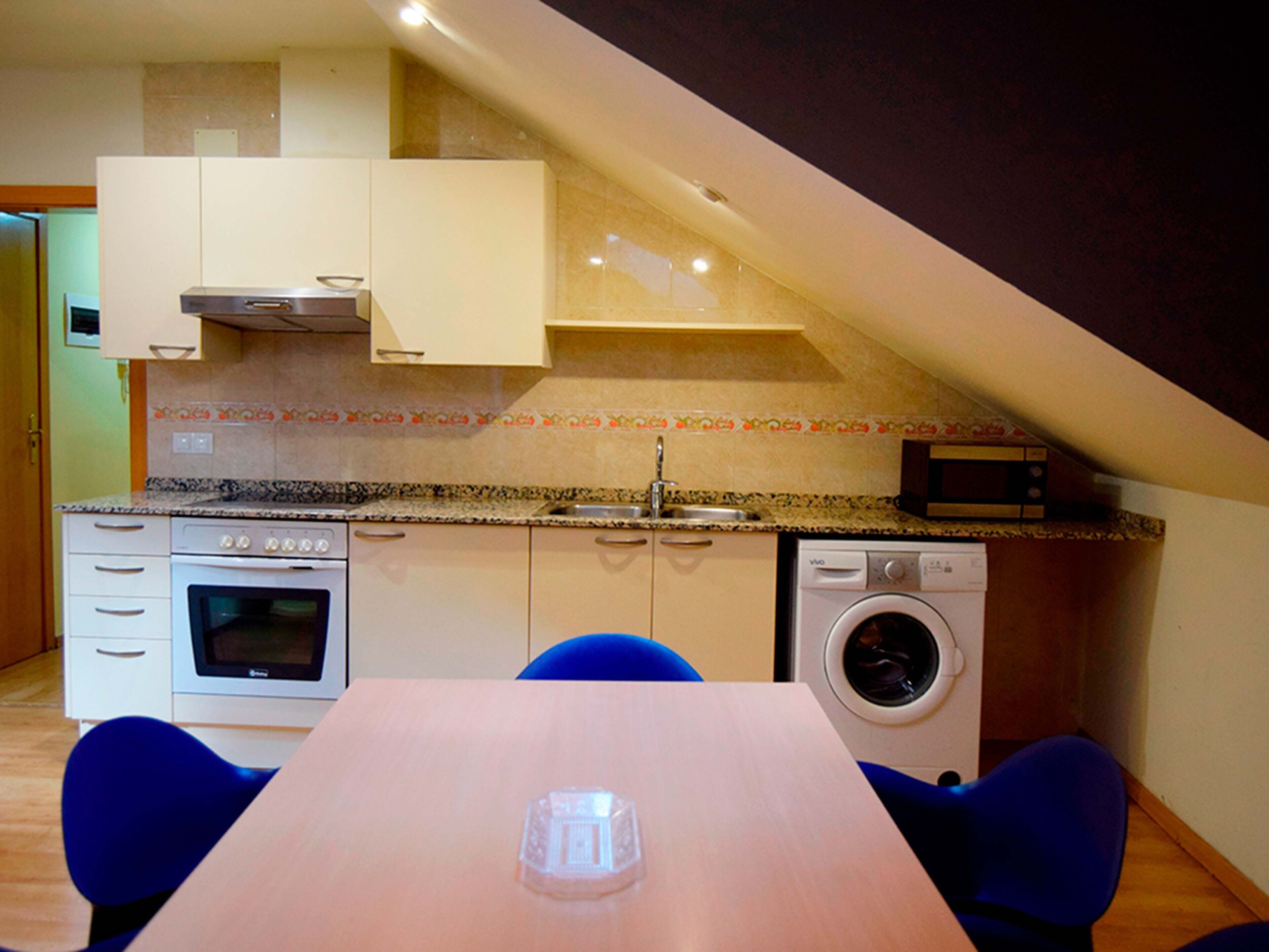 A compact kitchen with a built-in oven, microwave, and washing machine, a dining table with chairs, and a sloped ceiling.