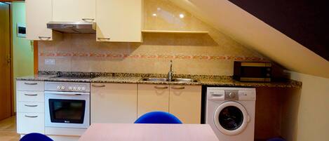 A compact kitchen with a built-in oven, microwave, and washing machine, a dining table with chairs, and a sloped ceiling.