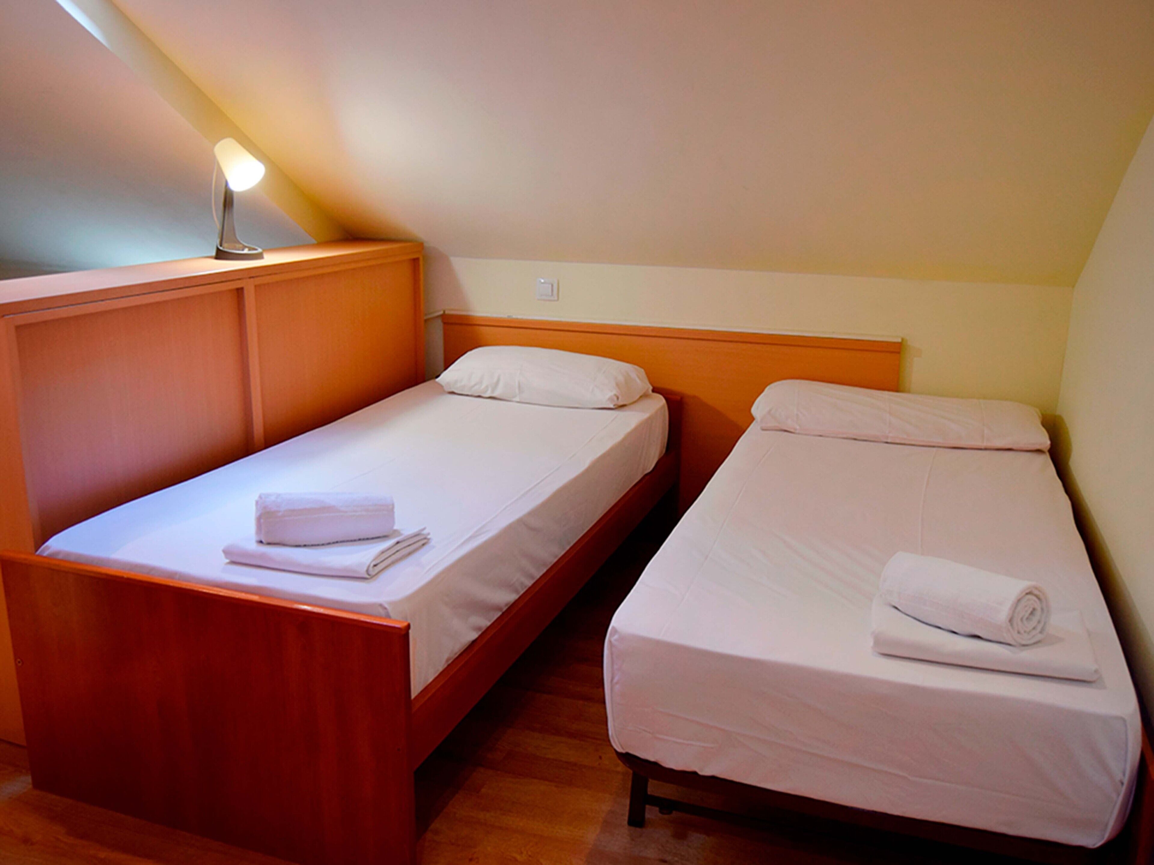 A room with two single beds, wooden headboards, white linens, and a bedside lamp.