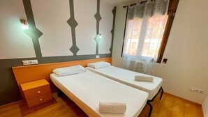 Une chambre avec deux lits simples, une table de chevet en bois et une fenêtre ornée d’un rideau à motifs floraux.