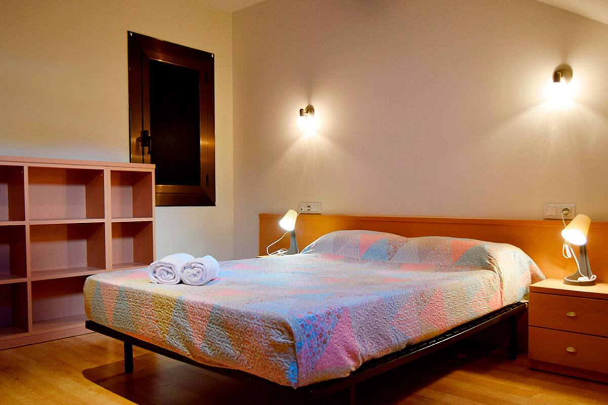 Ein ordentlich bezogenes Bett mit farbenfroher Steudecke, zwei Nachttischlampen, einem Holz-Kopfteil und einem Nachttisch mit Schublade.