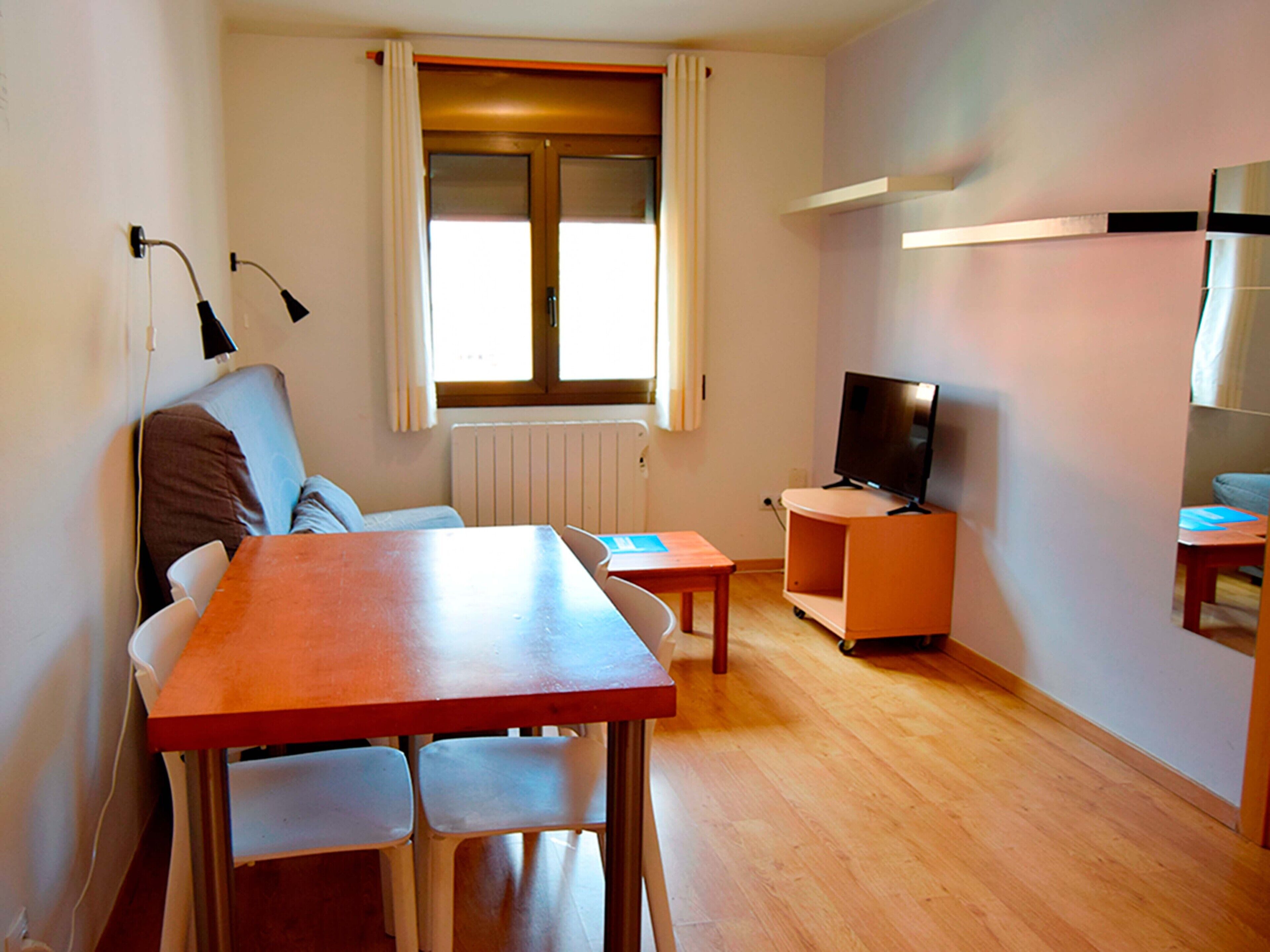 A compact living space with a wooden dining table, two chairs, a sofa, a television on a stand, and a window with curtains.