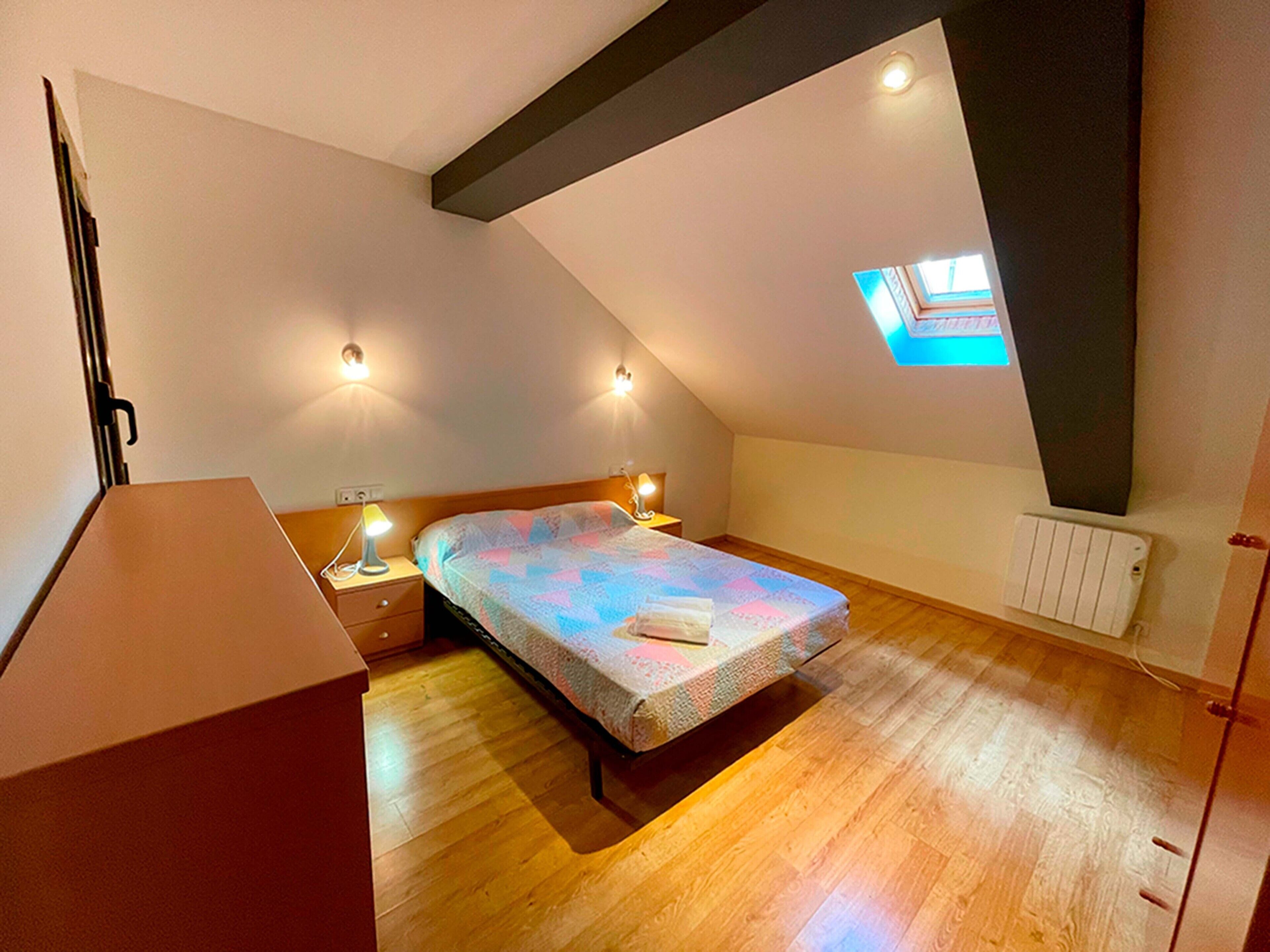 A bedroom with a sloped ceiling, a skylight, a bed with a quilt, bedside lamps, and a radiator.