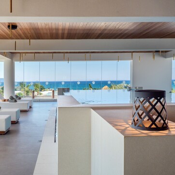 A modern lounge area with a pool, large windows offering a view of the sea, and a wooden ceiling.