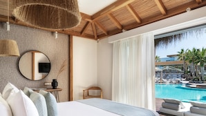 Un dormitorio con una cama grande, un espejo redondo, una mesita de noche con un florero, y vistas a una piscina y zona de descanso.