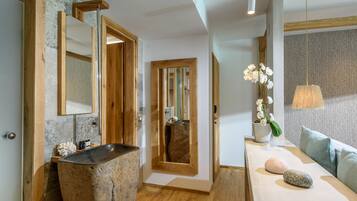 A modern bathroom with a large mirror, wooden accents, a stone sink, and a seating area with a plant.