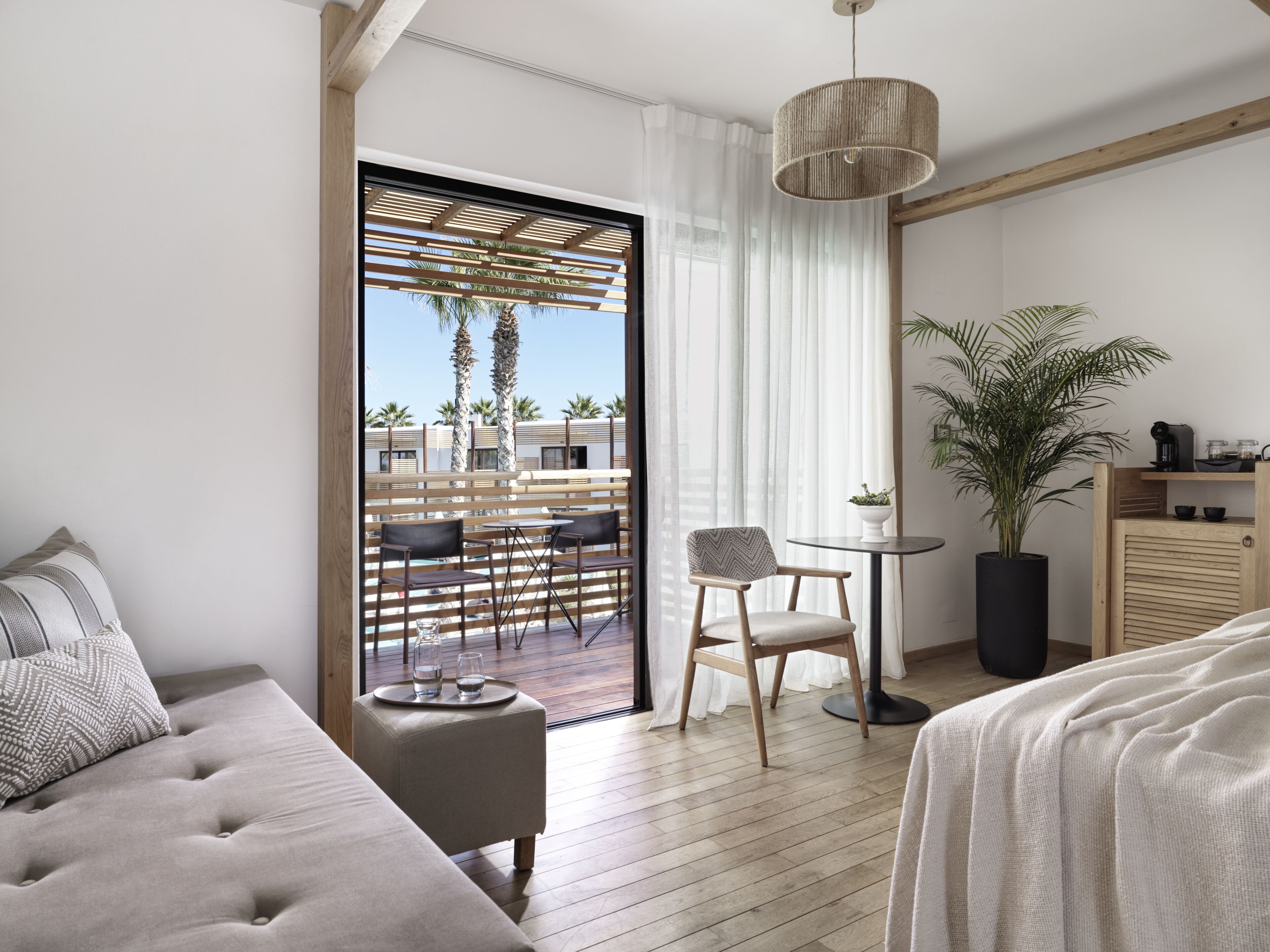 A modern hotel room with a balcony, a sofa, a chair, a small table, and a potted plant.