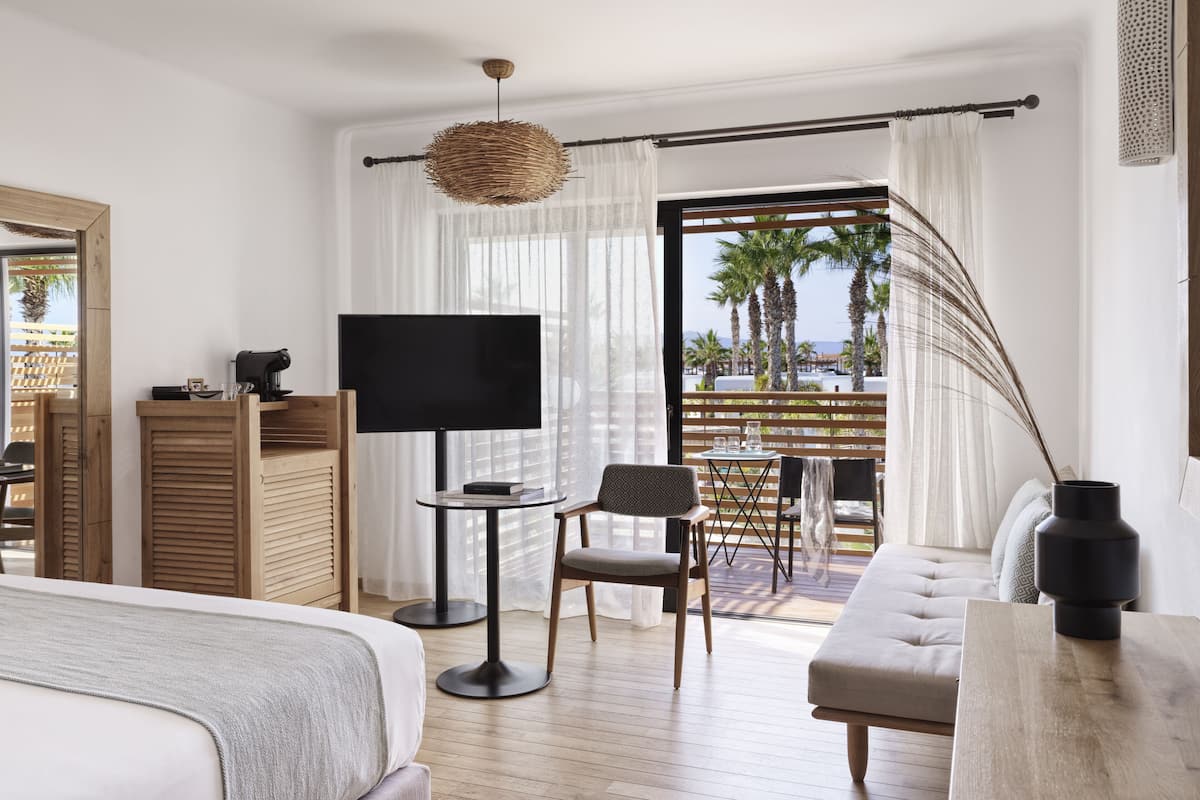 A modern hotel room with a large bed, a TV, a small table, and a balcony with a view of palm trees and the sea.