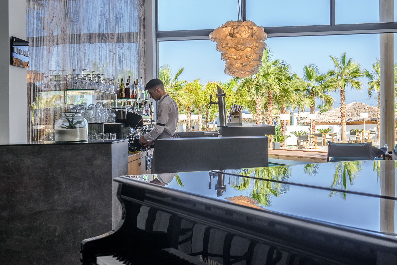 A bar area with a bartender, a piano, and a view of palm trees and a pool outside.