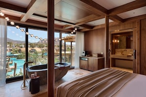 A modern hotel room with a large bed, a bathtub, a view of a pool and mountains, and a wooden interior.