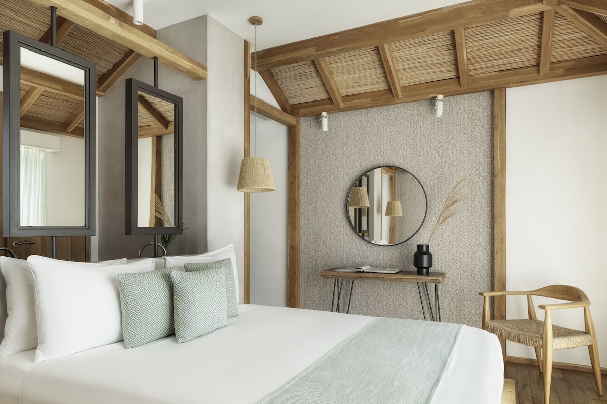 A modern bedroom with a large bed, wooden beams, a round mirror, a small table with a lamp, and a wicker chair.