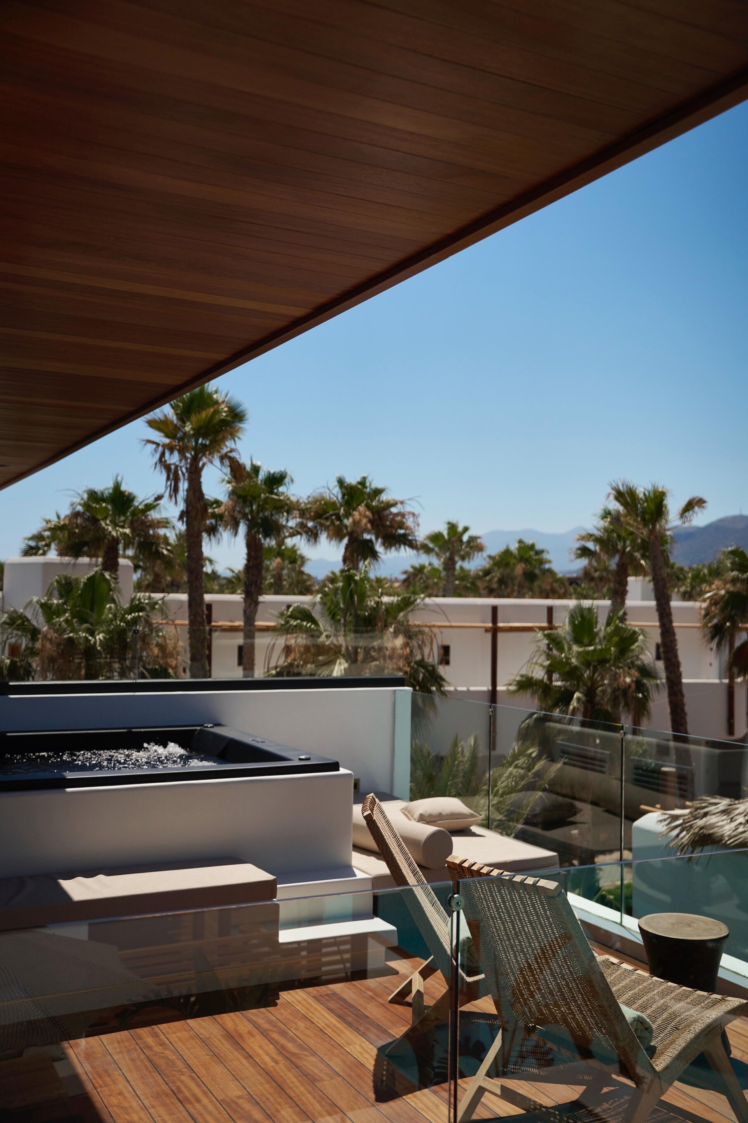 A modern outdoor lounge area with a hot tub, lounge chairs, and a glass balustrade overlooking palm trees and mountains.