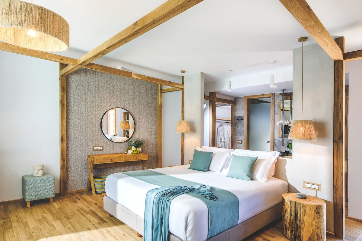 A bedroom with a bed, wooden beams, a mirror, bedside lamps, and a small stool.