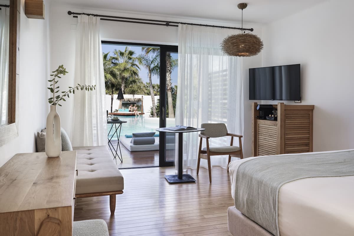 A hotel room with a bed, a TV, a dining table, a chair, a small pool, and a view of palm trees.