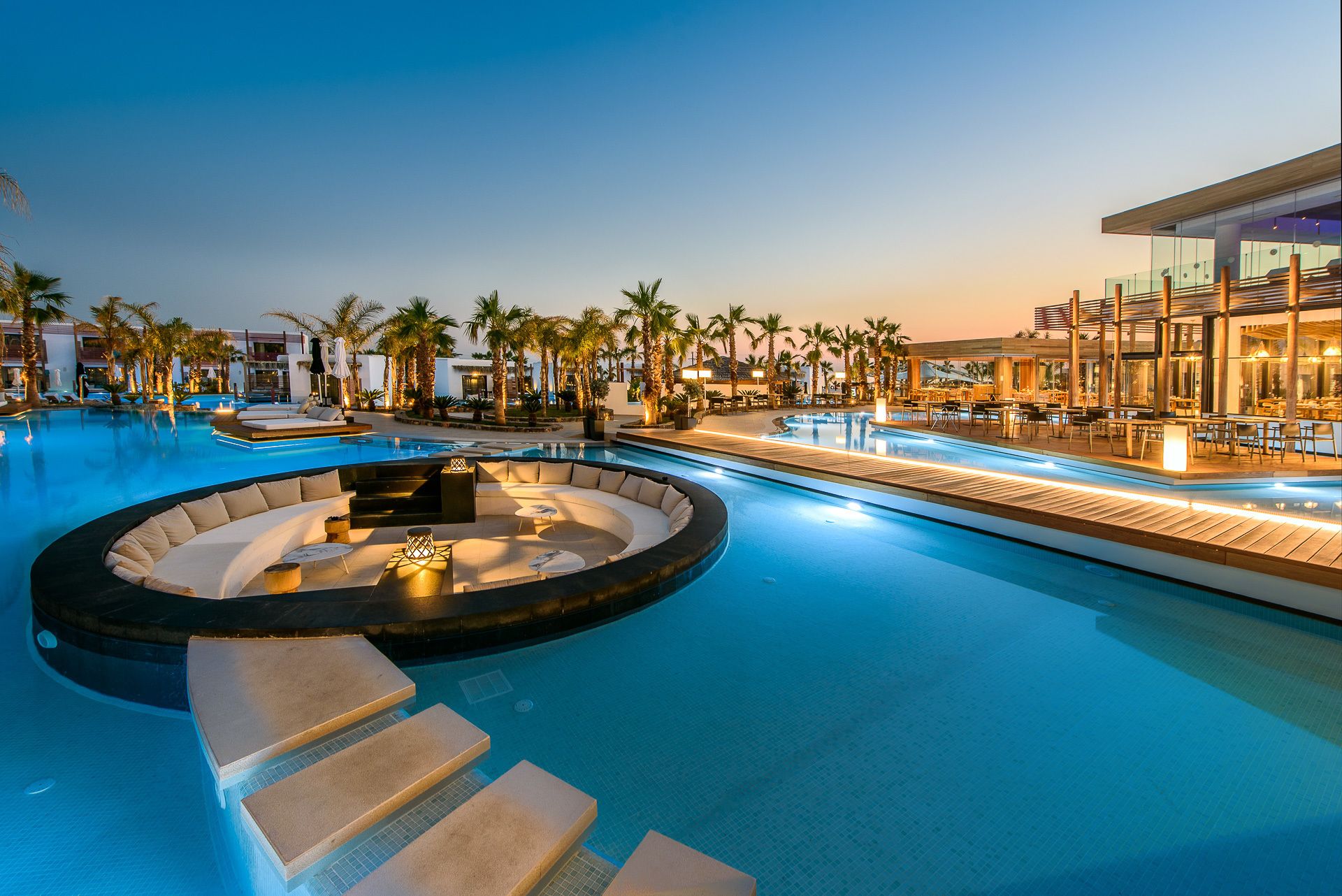 A pool area with a circular seating area, lounge chairs, and a restaurant with outdoor seating.