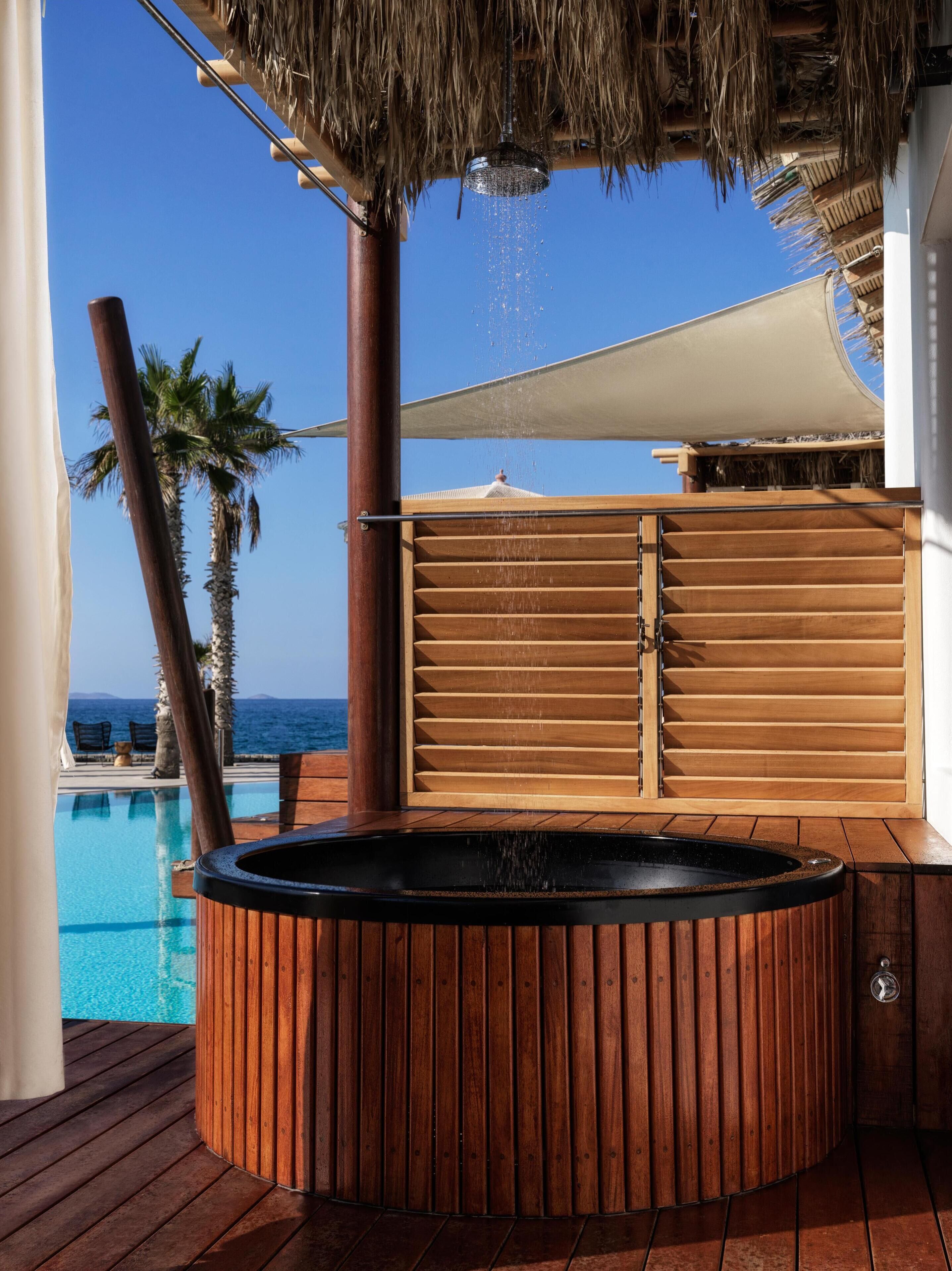 A wooden hot tub with a view of the ocean, a palm tree, and a pool.