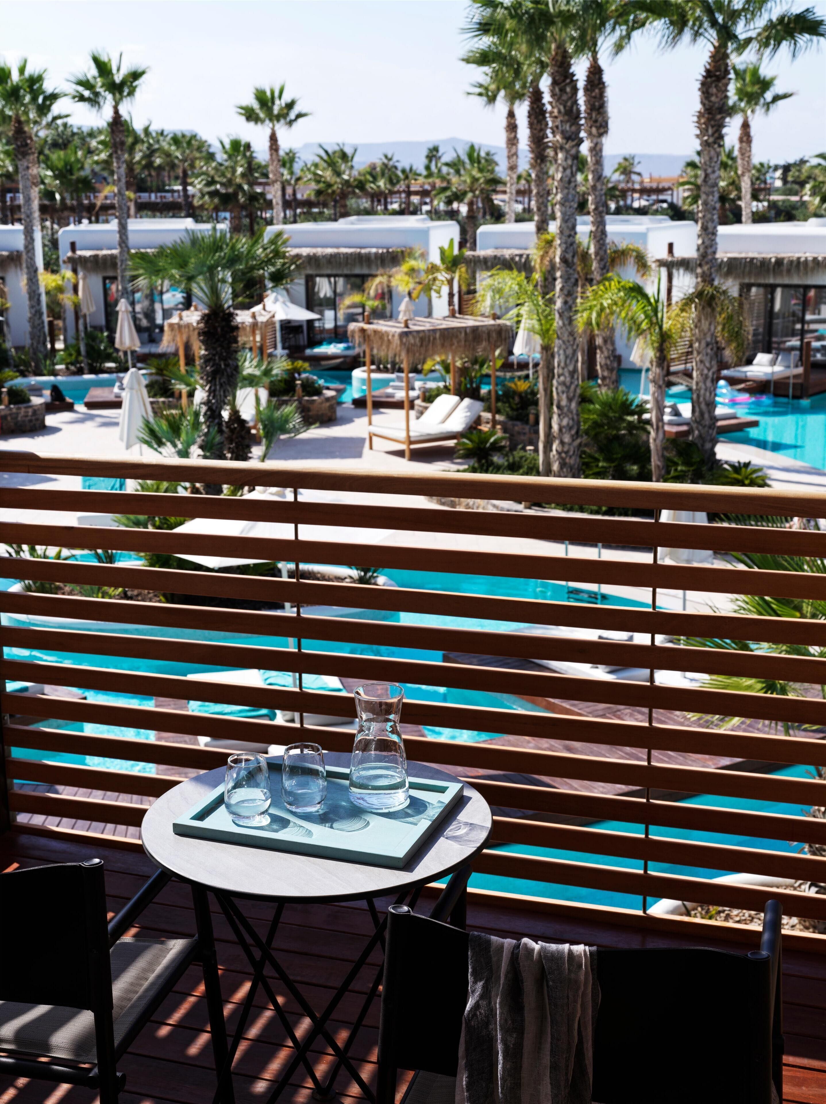 A poolside area with wooden slatted seating, a table with two glasses and a pitcher, and a view of a pool surrounded by palm trees and lounge chairs.