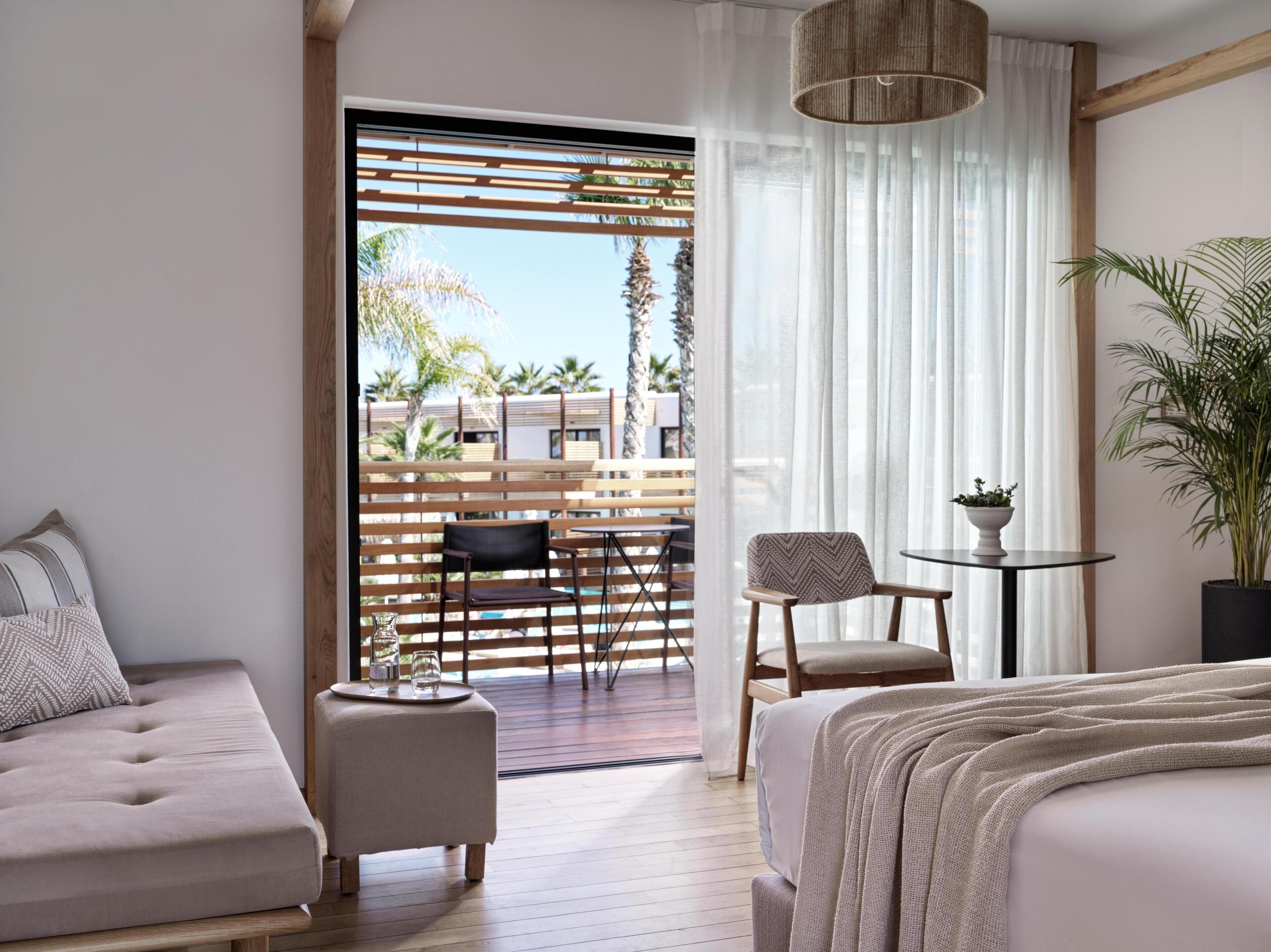 A modern hotel room with a large bed, a sofa, a small table with a plant, and a view of an outdoor patio with wooden furniture and a pool.