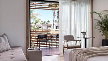A modern hotel room with a large bed, a sofa, a small table with a plant, and a view of an outdoor patio with wooden furniture and a pool.