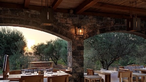 An outdoor dining area with stone arches, wooden furniture, and a view of olive trees.