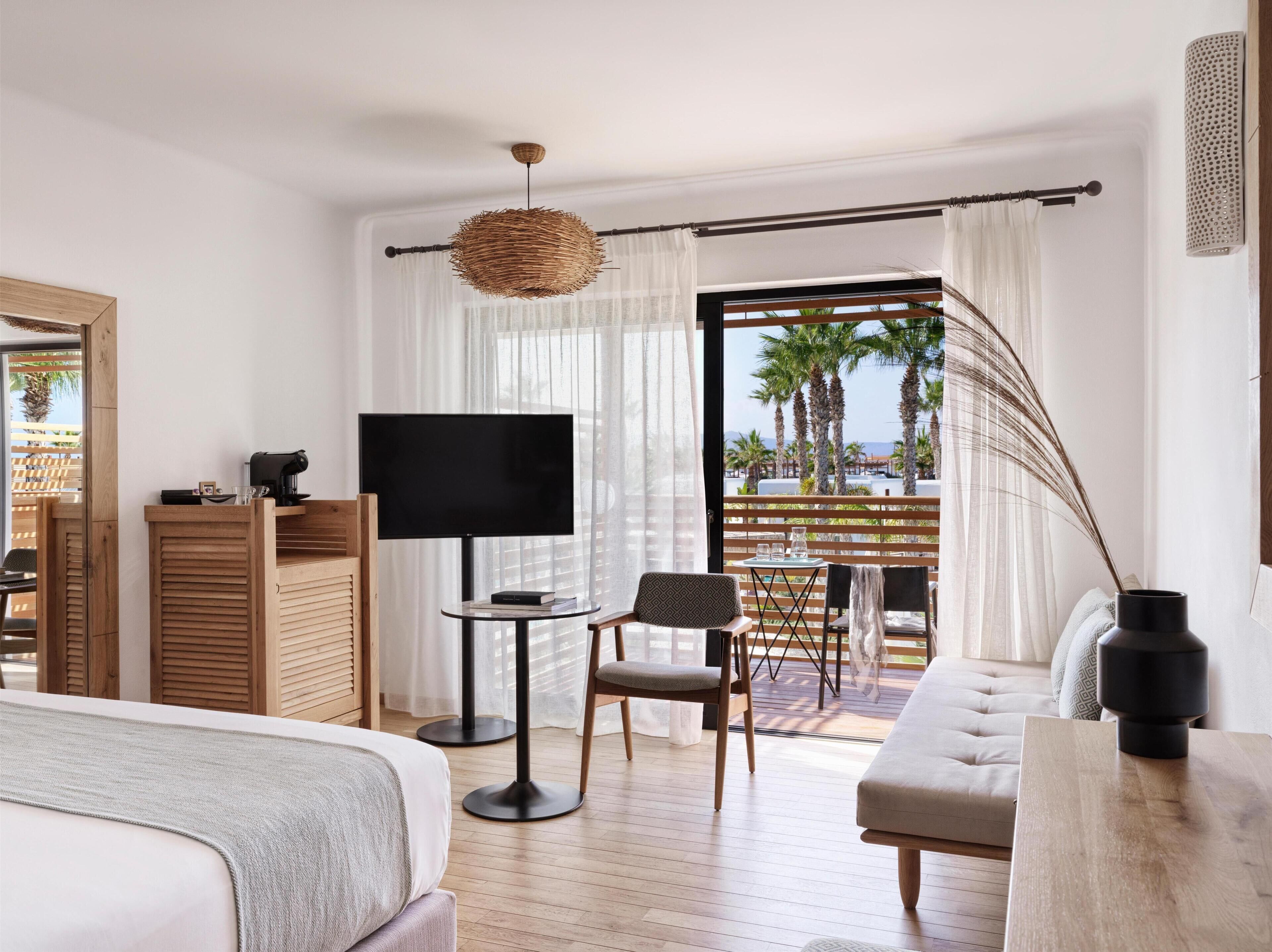 Une chambre d’hôtel comprenant un lit, une télévision, une petite table avec une chaise, un canapé et un balcon offrant une vue sur les palmiers et la mer.