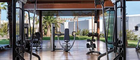 An open gym with various exercise equipment, a wooden floor, and a view of a white building and greenery outside.