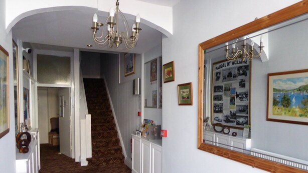A hallway with patterned carpet, a staircase, a chandelier, framed pictures, and a mirror reflecting a scenic painting. - The Berwick - Over 40's Only (Blackpool)