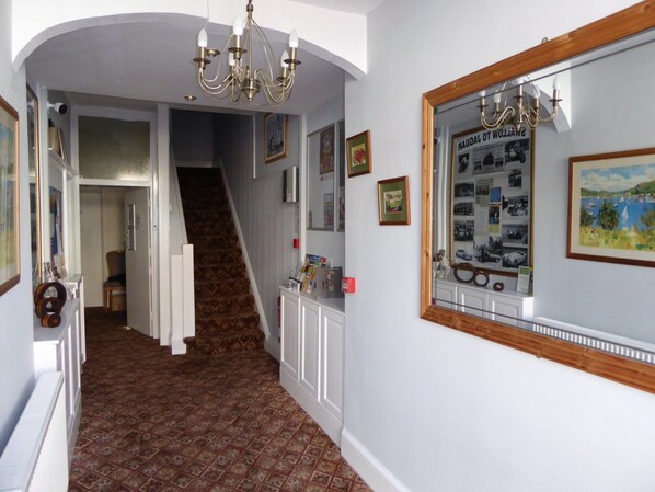 Un pasillo con alfombra estampada, una escalera, un candelabro, cuadros enmarcados y un espejo que refleja una pintura de paisaje.