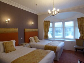 A hotel room with two beds, a chandelier, a window with curtains, and a chair. - The Berwick - Over 40's Only (Blackpool)