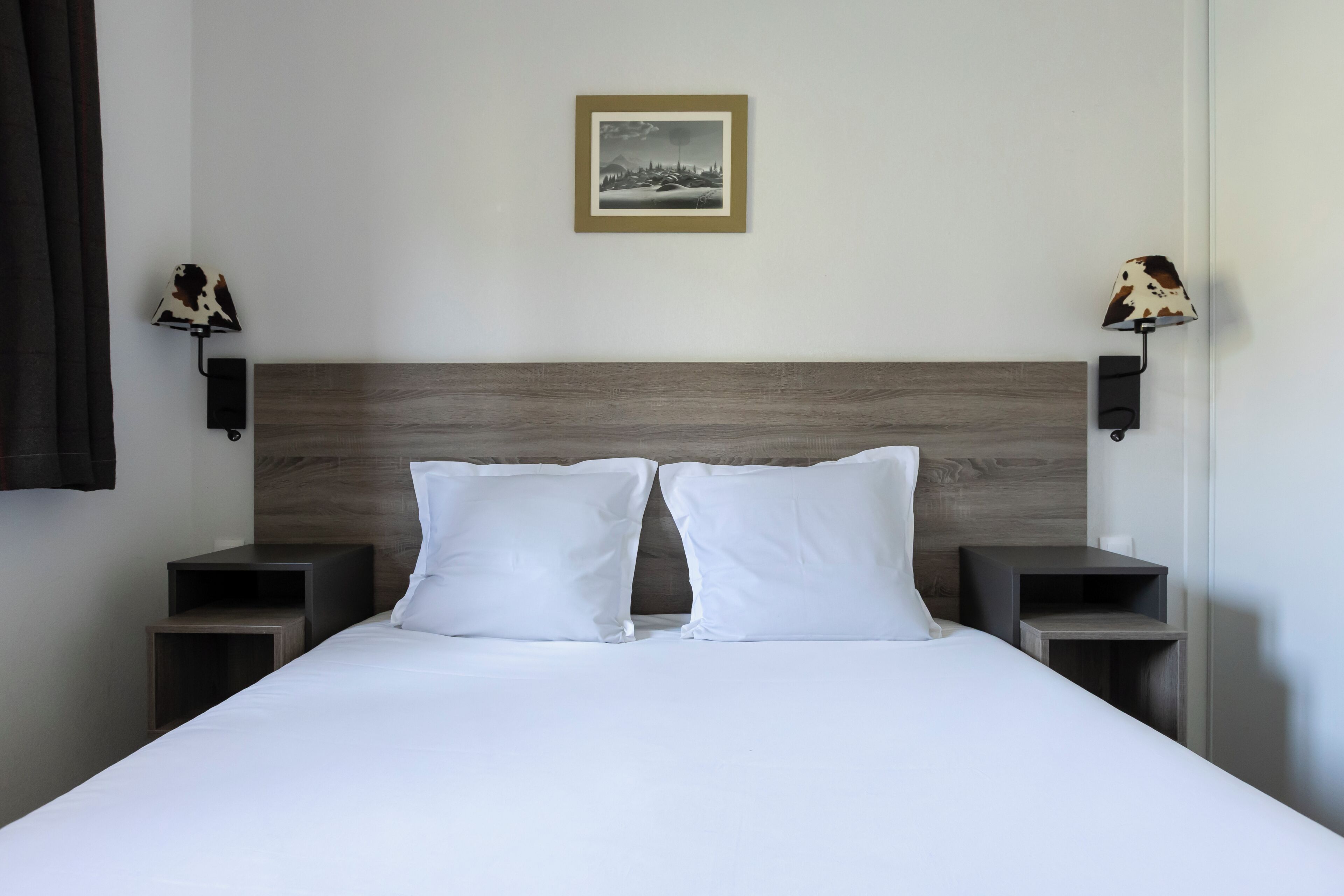 A neatly made bed with white linens, two bedside tables, a headboard, and two wall-mounted lamps with cowhide shades.